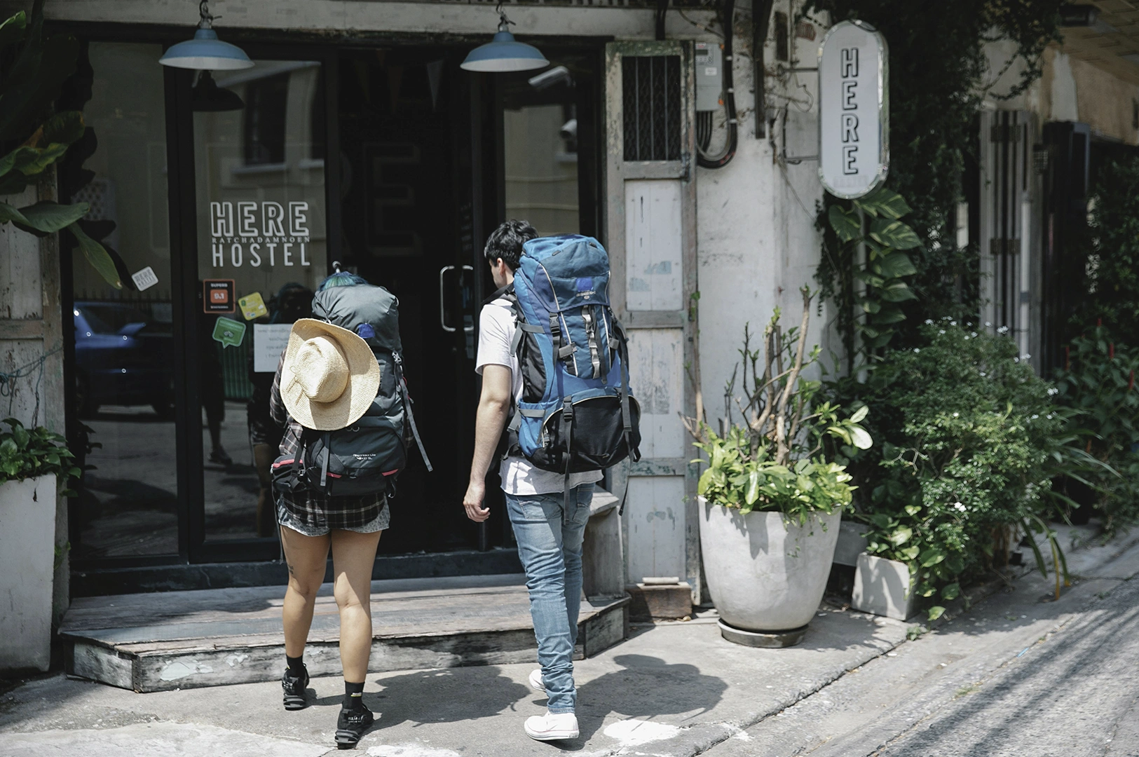 A high-angle, cinematic shot capturing two backpackers from behind as they approach "HERE Hostel." The woman on the left wears a plaid shirt, short shorts, and a wide-brimmed straw hat hanging over her large dark-grey trekking pack. The man on the right wears a white t-shirt, blue jeans, and a massive blue and black hiking backpack. They are walking toward a black-framed glass door of a rustic, white-walled building adorned with climbing green vines, a minimalist oval "HERE" sign, and potted plants. The lighting is bright and natural, suggesting a warm, sunny afternoon in an urban Southeast Asian setting.