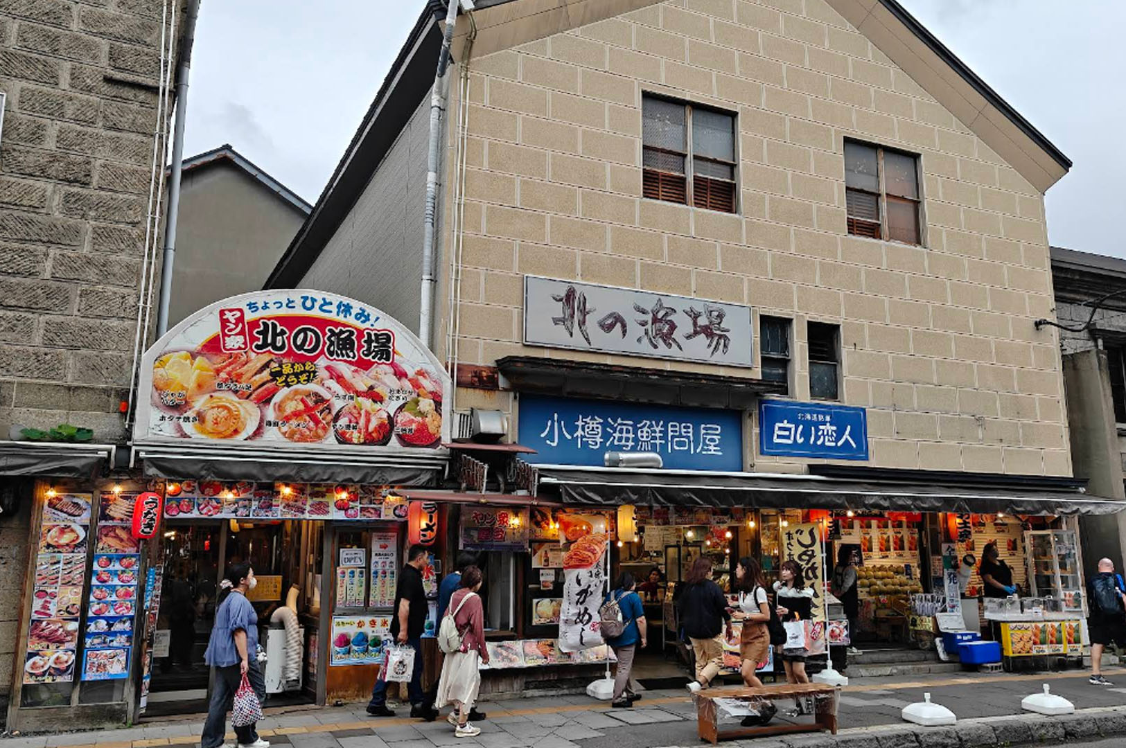 A street-level view of a bustling commercial building in Otaru. The main shop, Kita no Ryoba, features a large semi-circular sign displaying vibrant photos of seafood bowls and crab. Below, several open-air stalls sell street food and souvenirs. Pedestrians walk along the sidewalk under a grey, overcast sky. A blue sign for Shiroi Koibito (famed Hokkaido chocolate) is visible on the right.
