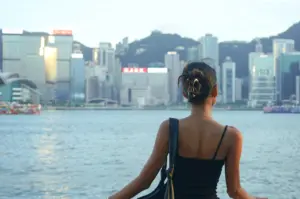 A medium shot from behind shows a woman with tan skin and dark hair in a messy bun secured by a gold claw clip, looking out over a wide body of blue water toward a dense city skyline nestled against rolling green hills. She wears a thin-strapped black top and a black shoulder bag with a knotted strap, with a delicate gold necklace resting against her upper back. In the background, the sun reflects brightly off the glass windows of various skyscrapers, while several traditional red-sailed and colorful boats dot the water's surface under a soft, clear sky.