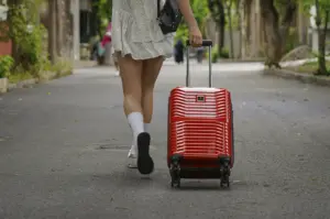 A low-angle, medium shot captured from behind shows a person walking down a quiet, paved street, pulling a vibrant, glossy red hardshell suitcase. The individual wears a white ruffled minidress with small black polka dots, white crew socks, and white sneakers, with one foot blurred in mid-stride. Their right hand grips the extended silver handle of the luggage, which features a textured, horizontal ribbed design. The background depicts a narrow, sunlit road lined with lush green trees and soft-focus buildings, creating a bright, airy atmosphere that evokes a sense of travel, new beginnings, or a morning arrival in a scenic neighborhood.