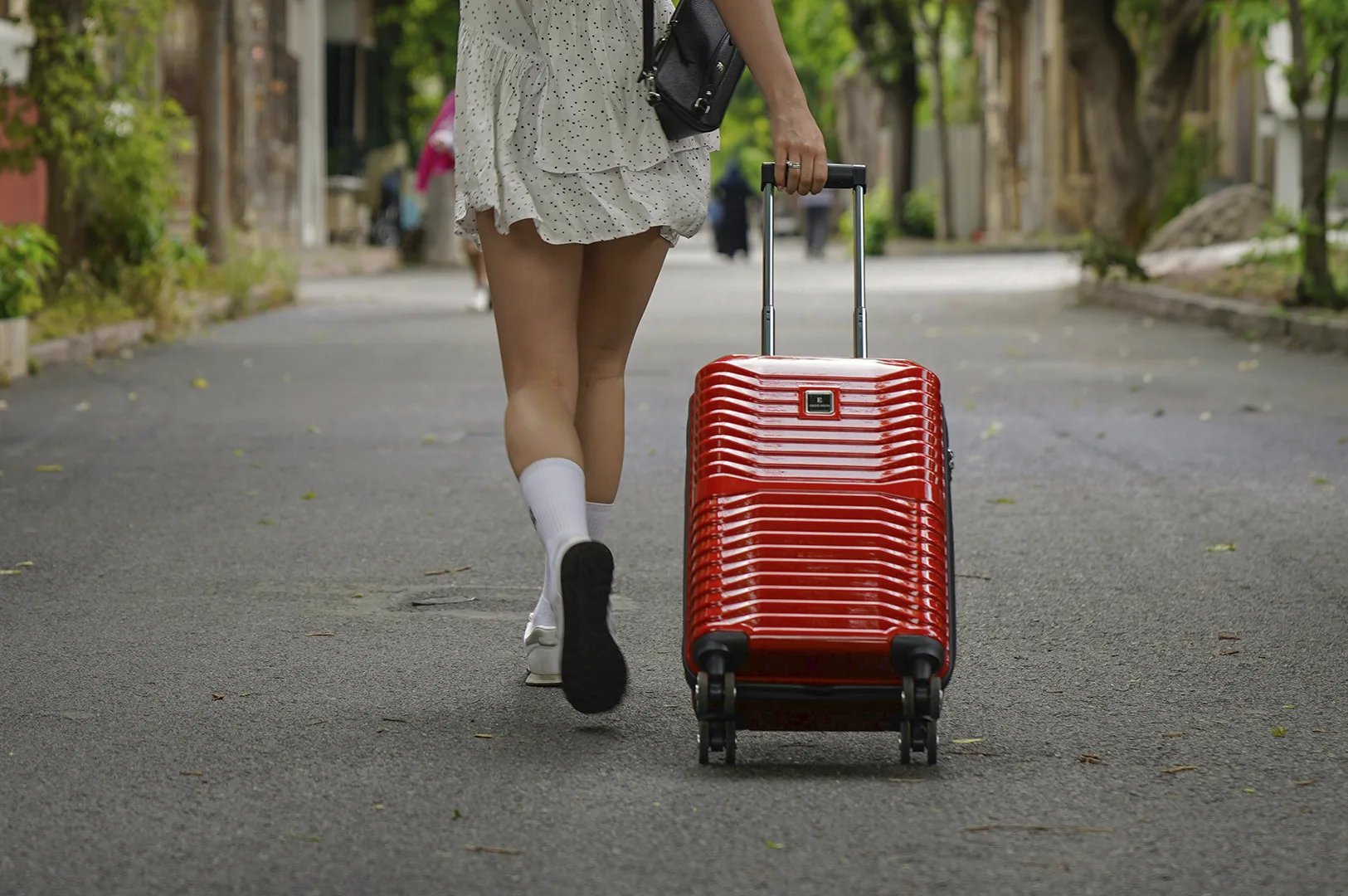 A low-angle, medium shot captured from behind shows a person walking down a quiet, paved street, pulling a vibrant, glossy red hardshell suitcase. The individual wears a white ruffled minidress with small black polka dots, white crew socks, and white sneakers, with one foot blurred in mid-stride. Their right hand grips the extended silver handle of the luggage, which features a textured, horizontal ribbed design. The background depicts a narrow, sunlit road lined with lush green trees and soft-focus buildings, creating a bright, airy atmosphere that evokes a sense of travel, new beginnings, or a morning arrival in a scenic neighborhood.