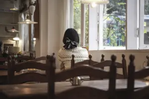 A serene, rear-view shot captures a person with dark hair tied in a checkered bow sitting alone at a wooden table in a quiet, rustic café. They are dressed in a cream-colored patterned sweater, looking out of a large, open window into a lush green garden bathed in soft, natural daylight. The interior is filled with warm, ambient light from vintage-style pendant lamps and features dark wood furniture, creating a lived-in and reflective atmosphere. The shallow depth of field blurs the foreground tables and chairs, drawing focus to the peaceful, solitary moment of "slow travel" and the tranquil neighborhood setting.