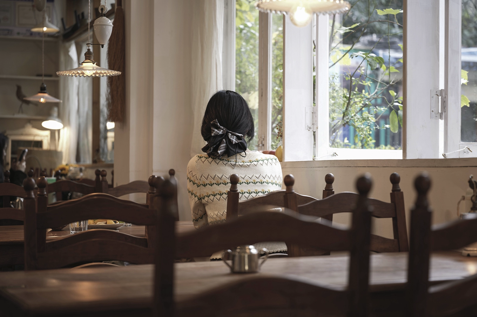 A serene, rear-view shot captures a person with dark hair tied in a checkered bow sitting alone at a wooden table in a quiet, rustic café. They are dressed in a cream-colored patterned sweater, looking out of a large, open window into a lush green garden bathed in soft, natural daylight. The interior is filled with warm, ambient light from vintage-style pendant lamps and features dark wood furniture, creating a lived-in and reflective atmosphere. The shallow depth of field blurs the foreground tables and chairs, drawing focus to the peaceful, solitary moment of "slow travel" and the tranquil neighborhood setting.