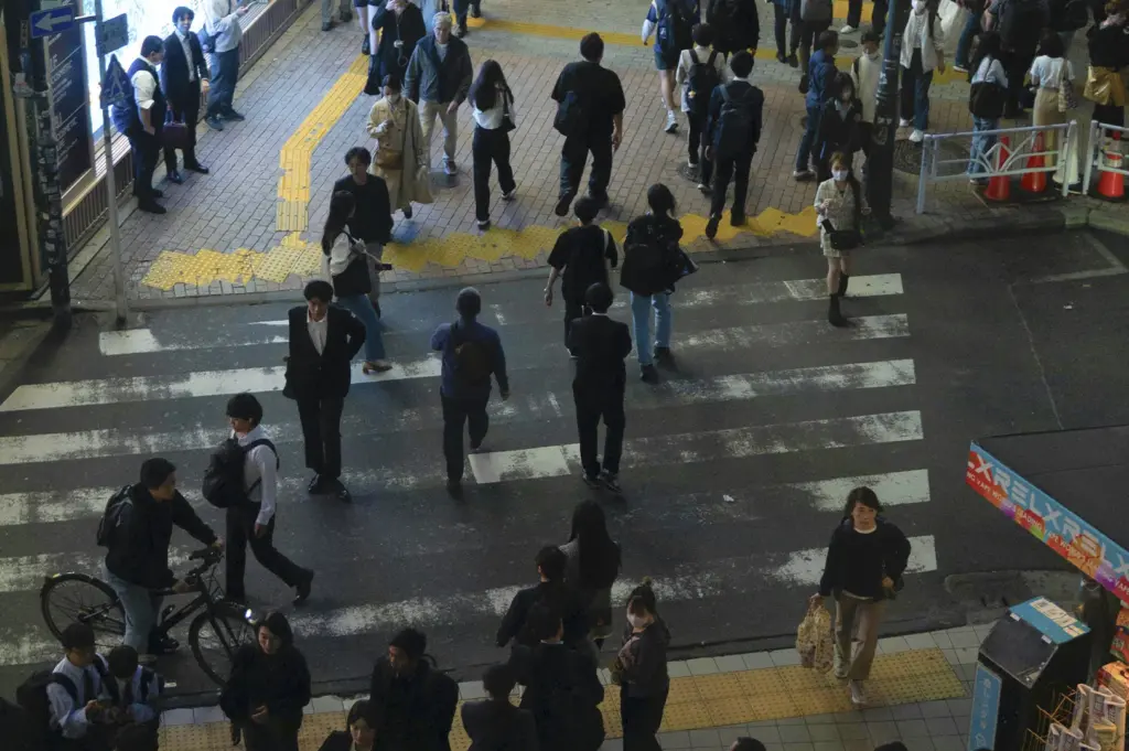 An elevated, high-angle shot captures a busy urban pedestrian crossing, likely in a Japanese city given the street markings and signage. Numerous people in professional and casual attire are mid-stride across the zebra-striped pavement, while a cyclist on the left waits to navigate through the crowd. The lighting is cool and slightly dim, suggesting late afternoon or an overcast day, highlighting the rhythmic, everyday motion of a bustling metropolitan hub.