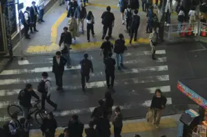 An elevated, high-angle shot captures a busy urban pedestrian crossing, likely in a Japanese city given the street markings and signage. Numerous people in professional and casual attire are mid-stride across the zebra-striped pavement, while a cyclist on the left waits to navigate through the crowd. The lighting is cool and slightly dim, suggesting late afternoon or an overcast day, highlighting the rhythmic, everyday motion of a bustling metropolitan hub.