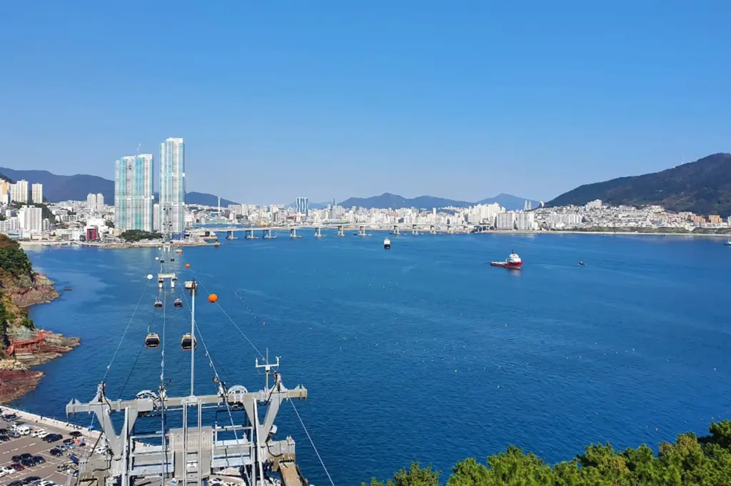 Overlooking the blue waters of Songdo Bay in Busan. In the foreground, the metal mechanical structure of a cable car station is visible, with several yellow and blue gondolas suspended on cables stretching across the sea. In the distance, the city skyline features tall modern skyscrapers and a long bridge connecting the coastal areas under a clear, bright blue sky.