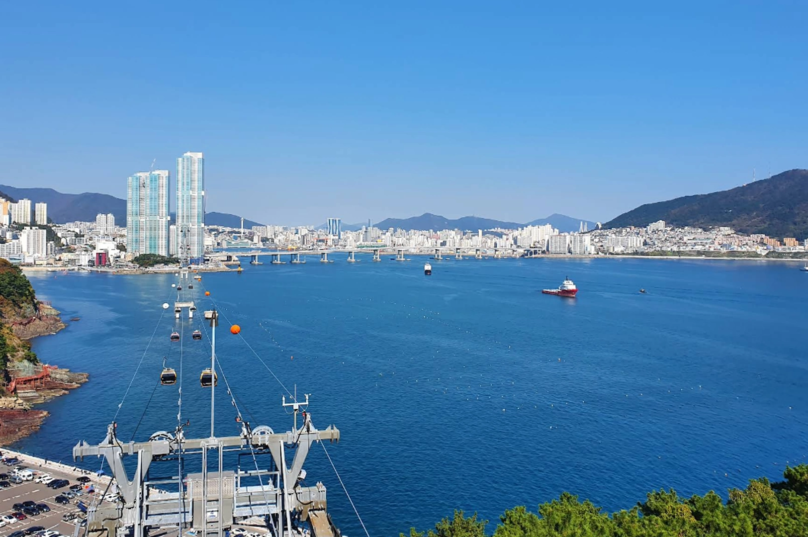 Overlooking the blue waters of Songdo Bay in Busan. In the foreground, the metal mechanical structure of a cable car station is visible, with several yellow and blue gondolas suspended on cables stretching across the sea. In the distance, the city skyline features tall modern skyscrapers and a long bridge connecting the coastal areas under a clear, bright blue sky.