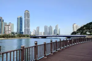 A wide-angle, eye-level shot from a wooden boardwalk looking across a calm blue bay toward the Marine City skyline in Busan. To the left, a modern blue bridge spans the water. In the background, several sleek, glass-paned skyscrapers and futuristic curved high-rises stand against a clear, pale blue sky. The wooden railing of the boardwalk runs along the bottom foreground, leading the viewer's eye into the coastal scene.