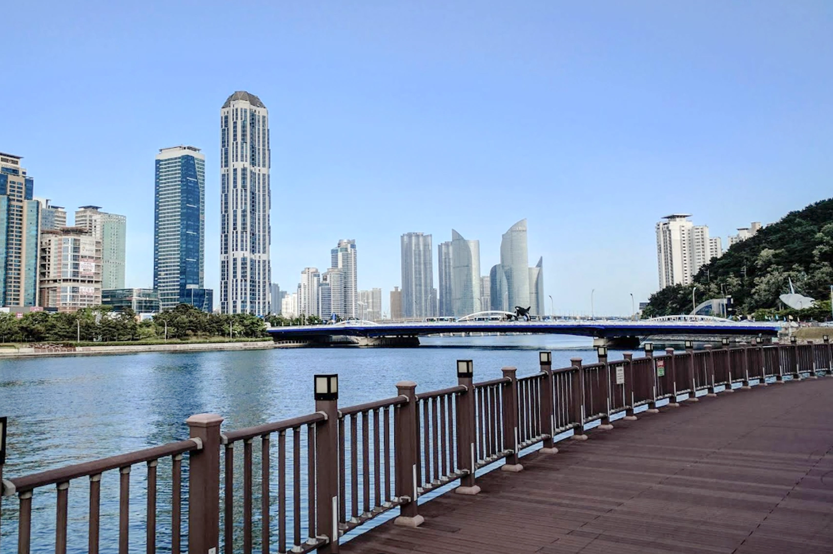 A wide-angle, eye-level shot from a wooden boardwalk looking across a calm blue bay toward the Marine City skyline in Busan. To the left, a modern blue bridge spans the water. In the background, several sleek, glass-paned skyscrapers and futuristic curved high-rises stand against a clear, pale blue sky. The wooden railing of the boardwalk runs along the bottom foreground, leading the viewer's eye into the coastal scene.