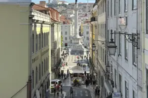 Full outdoor shot shows a cobbled stone street leading back to the middle background where a white marble monument stands, a pillar with a human figure at the top and a square, multi-tiered base, visible in full, in an old European city. On the left side, two to four-story yellow and grey stucco buildings run front to back; on the right side are three to four-story white buildings. The square base of the monument is directly in front of and in the bottom foreground is a cafe patio filled with red table cloths and umbrellas and about eight to twelve patrons sitting and dining. Many tourists are walking around, facing different directions and in small clusters, mostly wearing colorful t-shirts, some with bags slung over their shoulder. Behind the patio on the left side of the street is a white van. The street is lined with multiple black, antique lamp posts. Small terracotta tiled roofs are visible behind and to the left of the monument. A light blue sky is visible at the very top. At the bottom of the street is patterned white and black tile, while the foreground is cobblestone. All sides of the street run front-to-back, giving a funnel shape, and are full of shadows but still brightly lit in a warm, summer sun. The monument pillar is detailed with multiple carvings of human figures.
