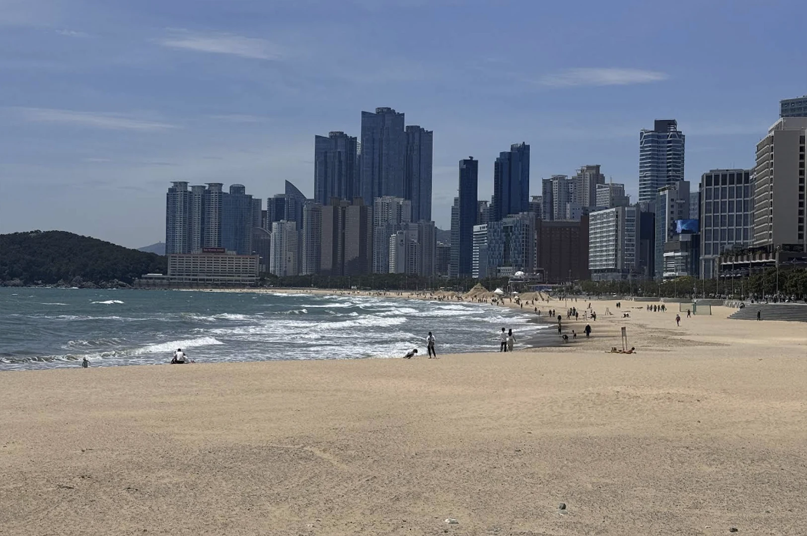 A wide, eye-level shot of a sprawling sandy beach meeting the crashing white surf of the ocean. In the distance, a massive cluster of modern, blue-glass skyscrapers and luxury apartments rises sharply against a hazy blue sky. Scattered groups of people are walking along the shoreline or sitting on the sand, emphasizing the scale of the urban coastline.
