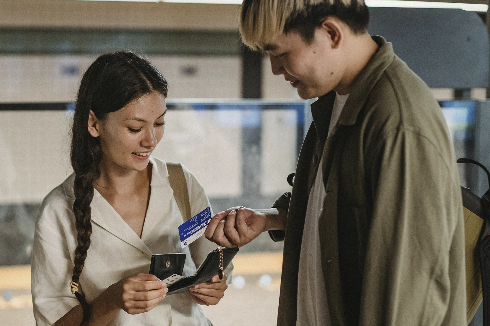 A medium shot of a young man and woman standing in a brightly lit, modern transit station. The man, with bleached blonde hair and a dark olive-green button-down shirt over a white tee, leans in slightly toward the woman. The woman, with a long dark braid and a cream-colored linen shirt, smiles warmly as she holds open a black leather wallet to retrieve a blue transit card. The background is softly blurred, showing the glass partitions and sleek architecture of a train platform, creating an atmosphere of shared excitement and casual travel.