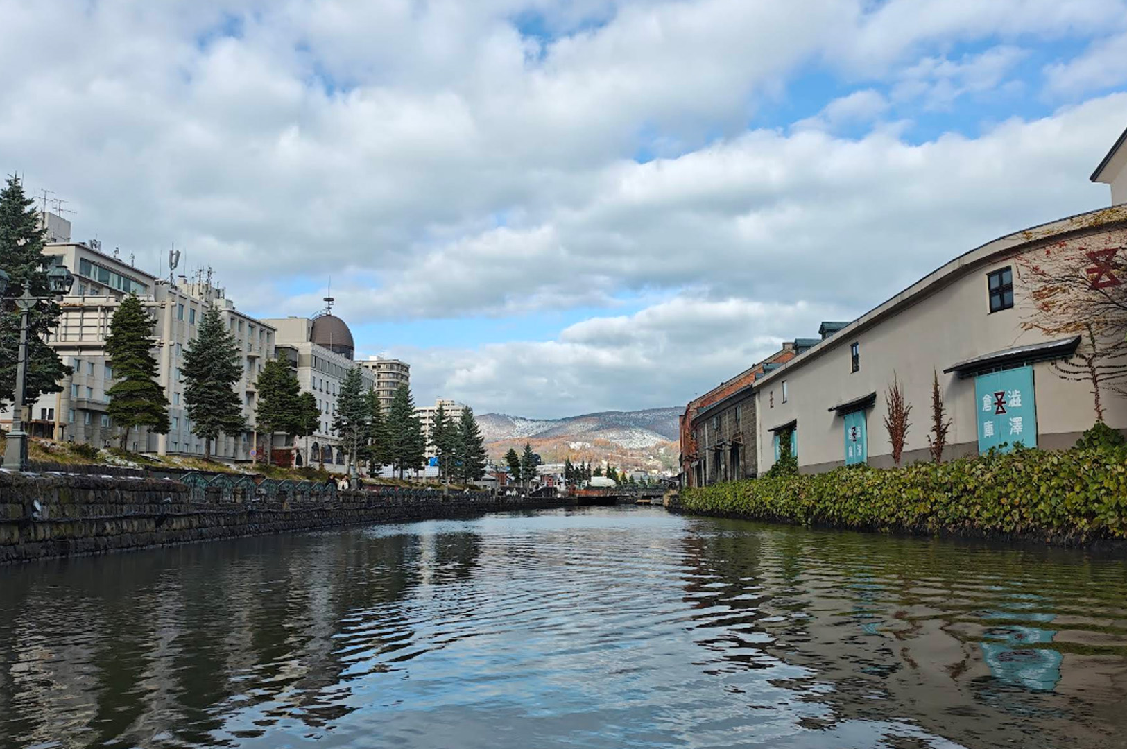 A wide, low-angle shot taken from the water of the Otaru Canal. To the right, historic stone and brick warehouses with white walls and green vines line the water's edge. To the left, a stone-paved walkway is bordered by manicured pine trees and modern hotel buildings. In the far distance, rolling mountains are lightly dusted with snow under a bright, cloudy sky.