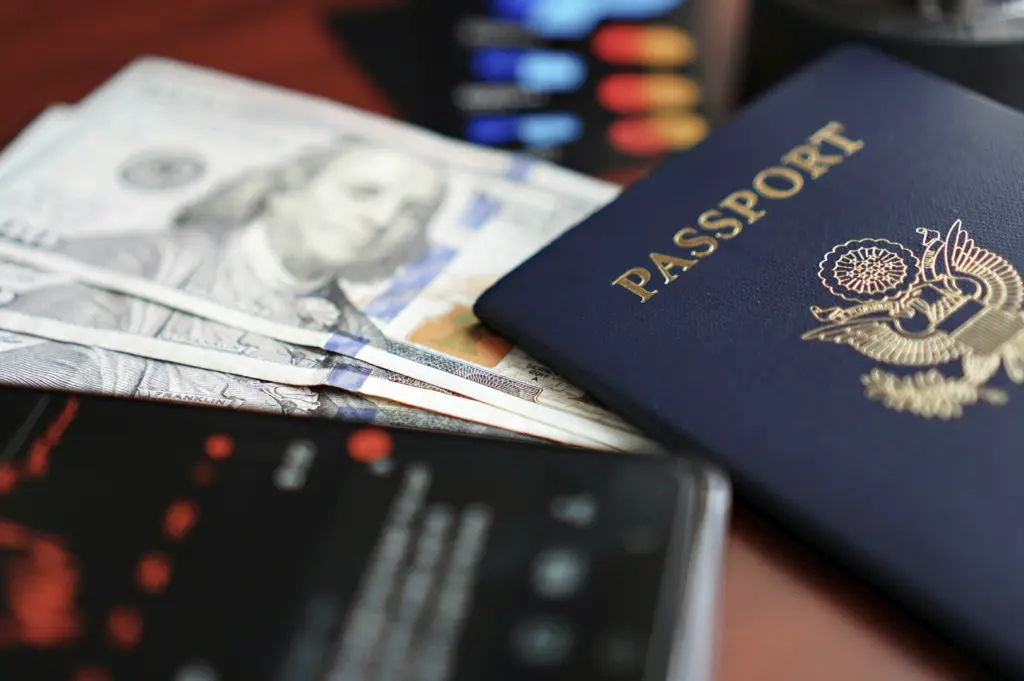A close-up, shallow-depth-of-field photograph features essential travel items resting on a dark surface. A blue United States passport with its gold-embossed eagle emblem is the primary focus on the right, partially overlapping several crisp $100 bills. In the foreground, the blurred screen of a smartphone displays what appears to be a financial or stock market tracking app with red and green data points, emphasizing the intersection of global travel and financial management.