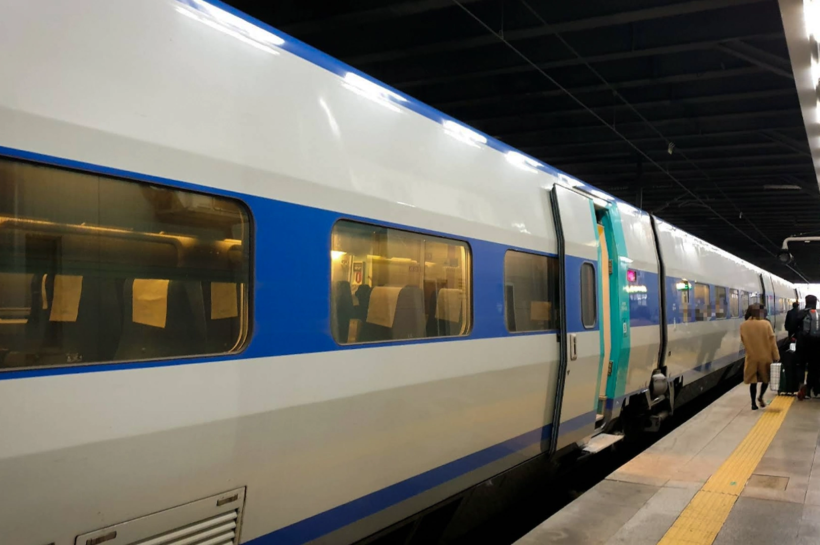 Modern KTX high-speed train stopped at a station platform at night. The train is white with a bold blue horizontal stripe and turquoise accents around the doors. Through the large rectangular windows, the warm yellow interior lighting reveals rows of passenger seats. A person in a tan coat walks along the platform next to the train.