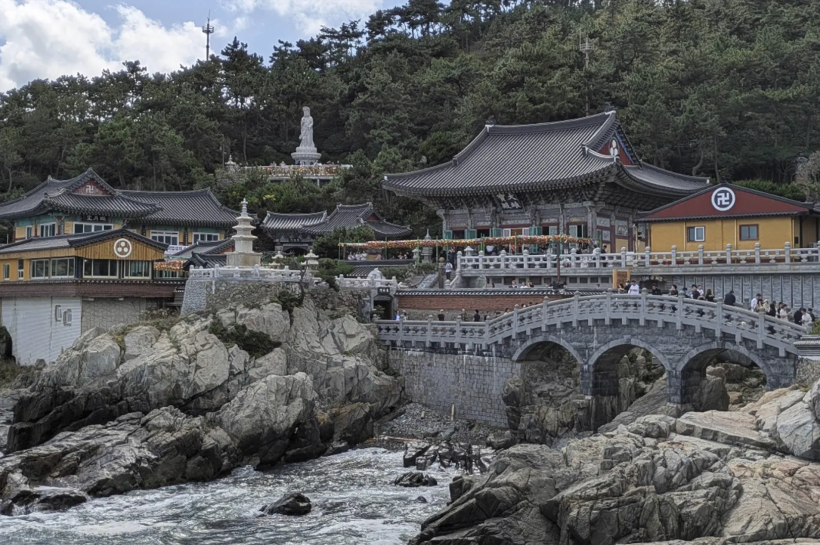 A scenic view of the Haedong Yonggungsa Temple, a traditional Korean Buddhist complex built directly onto rugged, rocky cliffs overlooking the sea. The architecture features dark tiled roofs with ornate colorful eaves and a prominent stone bridge with three arches. A tall white stone statue of a deity stands on a higher terrace amidst lush green pine trees, while waves crash against the jagged rocks in the foreground.
