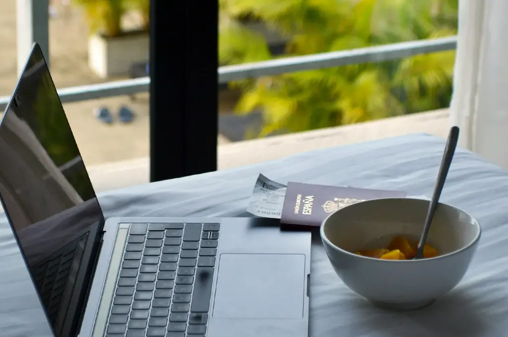 A close-up, high-angle shot captures a travel planning setup on a white bedspread near a window with a view of tropical greenery. A sleek, silver laptop is open on the left, while a dark red European Union passport with "España" embossed on the cover sits in the center next to a printed airline boarding pass. To the right, a white ceramic bowl contains pieces of fresh yellow fruit with a metal spoon resting inside, suggesting a relaxed morning spent organizing travel details from a hotel or vacation rental.