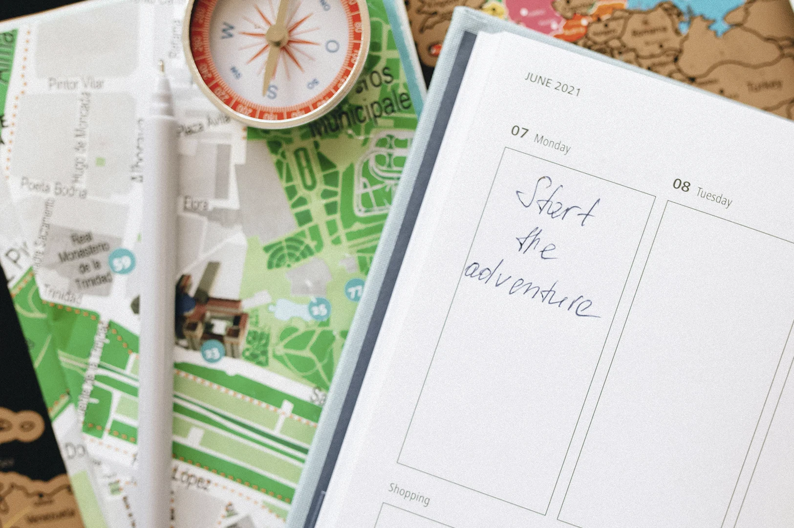 A top-down, flat-lay macro photograph focused on the tools of travel planning. On the right, an open white planner displays the date "JUNE 2021" with the handwritten phrase "Start the adventure" in blue ink under Monday the 7th. To the left, a vibrant green and white city map is partially visible, topped with a white stylus pen and a classic analog compass with a red and silver casing. The composition is clean and organized, evoking a sense of anticipation, precision, and the romanticism of traditional navigation.