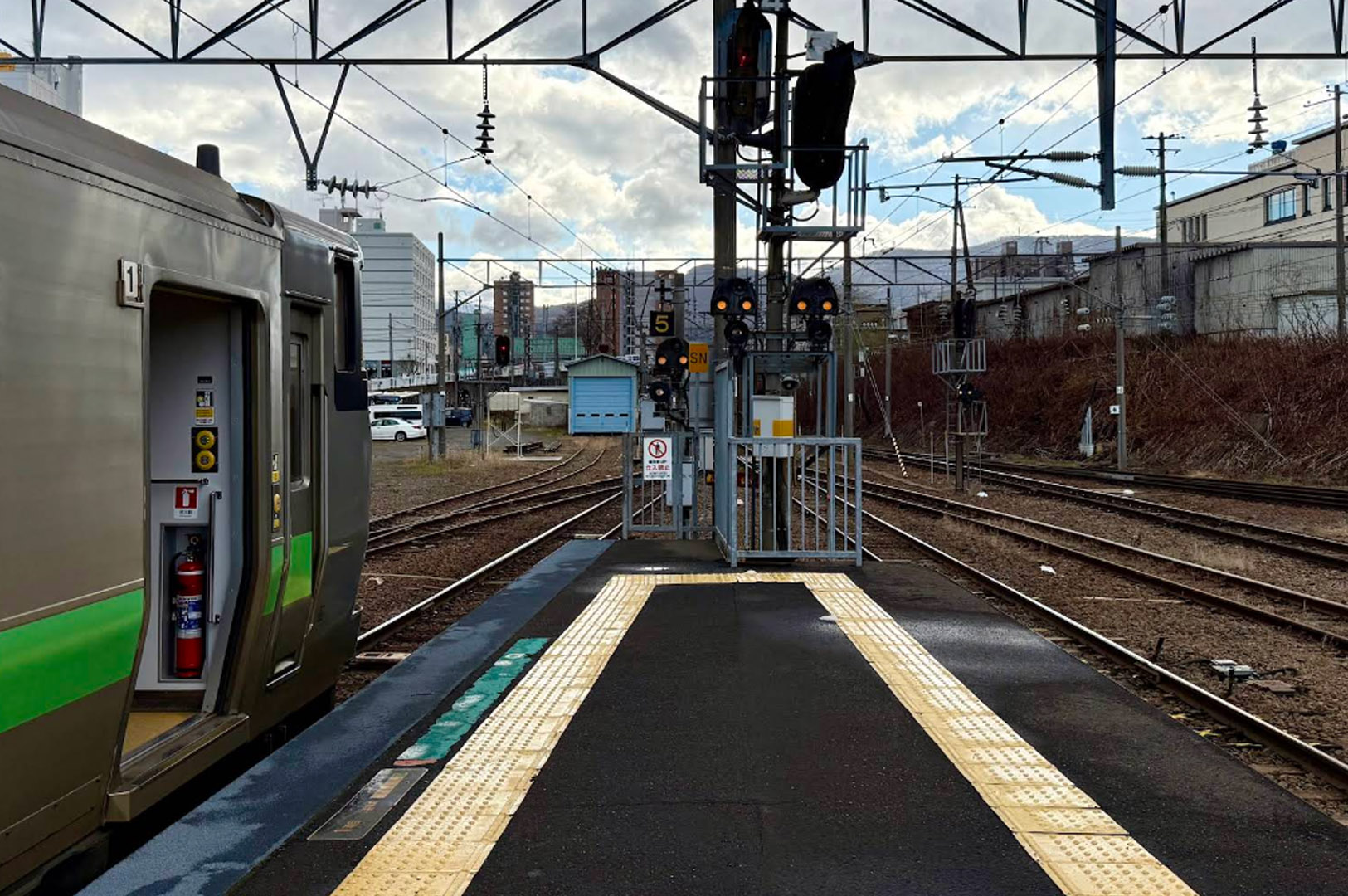 A perspective shot from a train platform looking down the tracks. On the left, a silver and green JR Hokkaido commuter train is stopped with its doors open, revealing a red fire extinguisher inside. The tracks curve into the distance toward a small blue maintenance shed. Overhead, a complex web of electrical wires and railway signals frames a backdrop of city buildings and mountains.