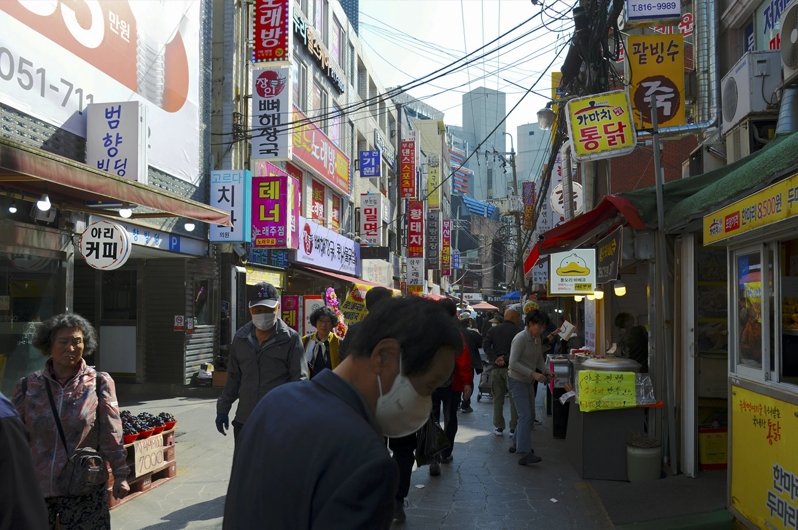 A busy, narrow street scene in a South Korean urban market area. The street is flanked by multi-story buildings covered in colorful vertical shop signs with Korean text (Hangul). Pedestrians, some wearing face masks, walk along the cracked pavement. On the right, small food stalls with yellow banners are visible, and on the left, a crate of dark grapes is for sale. A complex web of power lines stretches across the sky.
