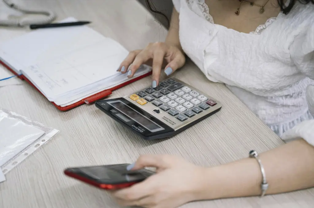 A top-down view shows a woman with light blue nail polish sitting at a light wood desk, actively managing finances. Her left hand holds a red smartphone, while her right hand operates a large Casio electronic desktop calculator. To her left lies a red-bound open planner with handwritten notes. She is wearing a white, textured lace blouse, and the overall composition suggests a moment of focused personal administration or trip planning.