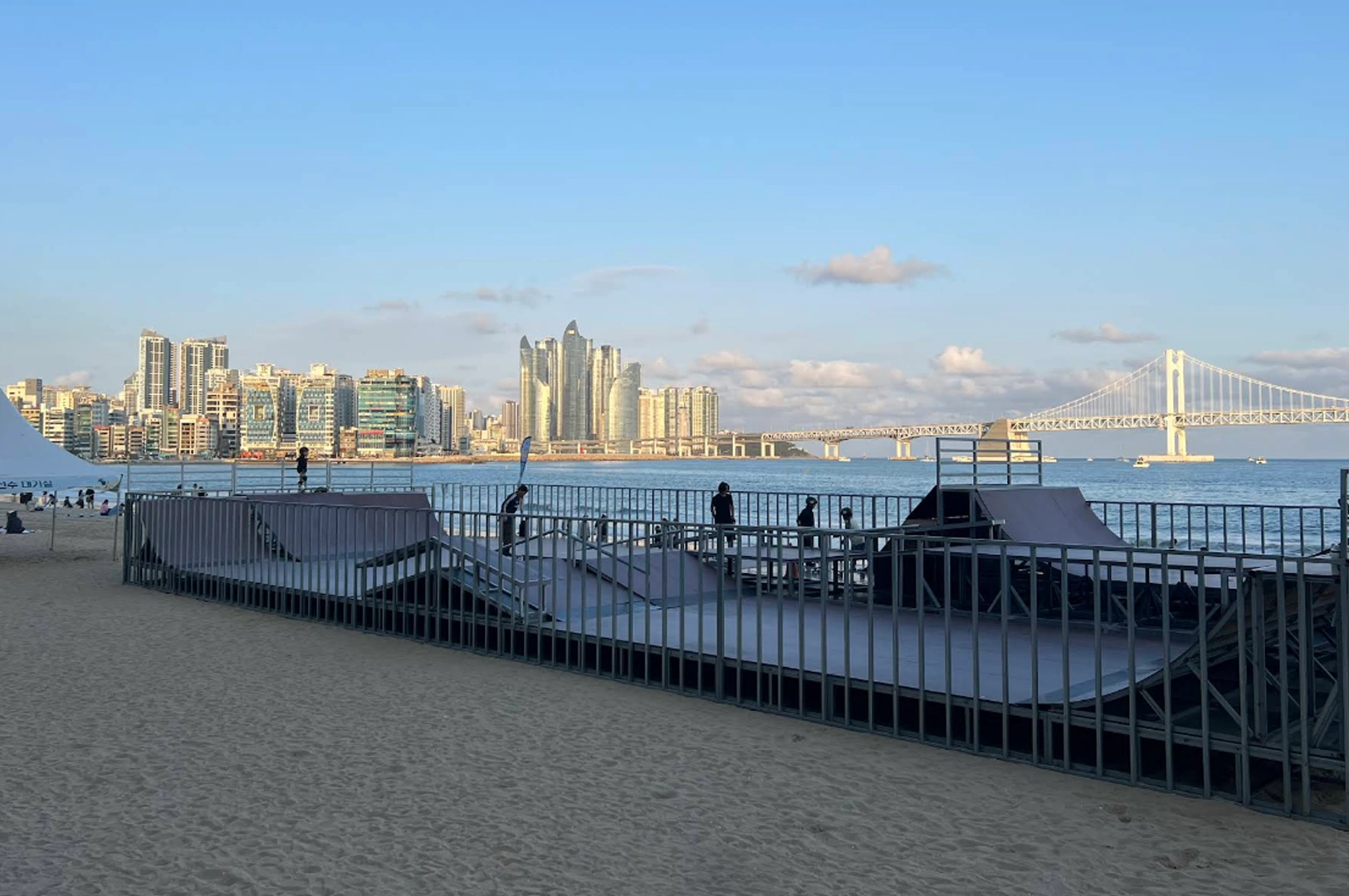 Gwangalli Beach at dusk. In the center foreground, there is a gray metal skate park ramp set on the sand. In the background, the calm ocean water leads to the Gwangandaegyo (Diamond Bridge), a massive white suspension bridge. The Busan city skyline, featuring a cluster of unique, curved skyscrapers at Marine City, sits under a pale blue sky with scattered light clouds.