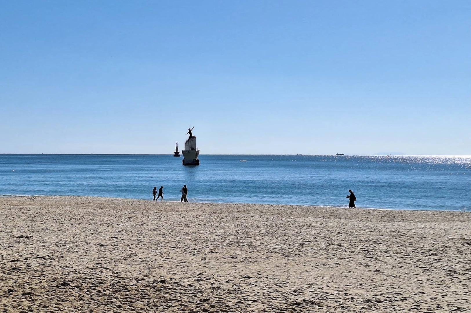 A bright, minimalist coastal shot of Haeundae Beach on a sunny day. The foreground is composed of textured, light-tan sand with a few distant silhouettes of people walking along the shoreline. The sparkling turquoise ocean occupies the mid-ground, featuring a unique white-and-red lighthouse structure shaped like a boat's hull positioned further out in the water. The horizon line is sharp against a cloudless, deep blue sky.