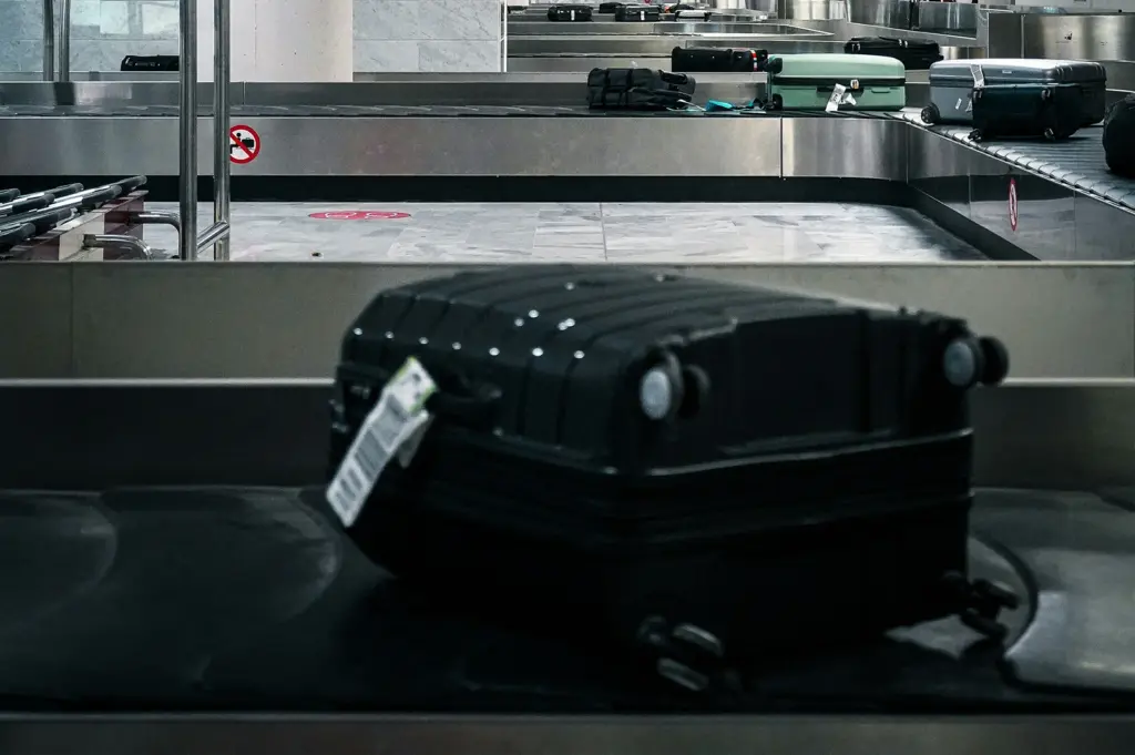 A black hardshell suitcase with a white baggage tag moves along a metal airport luggage carousel. The focus is sharp on the suitcase in the foreground, while other bags, including a teal suitcase and several black ones, are visible in the soft-focus background on a parallel conveyor belt. The scene is set in a modern, brightly lit airport baggage claim area with grey tiled floors and a "no sitting" sign visible on the side of the carousel.