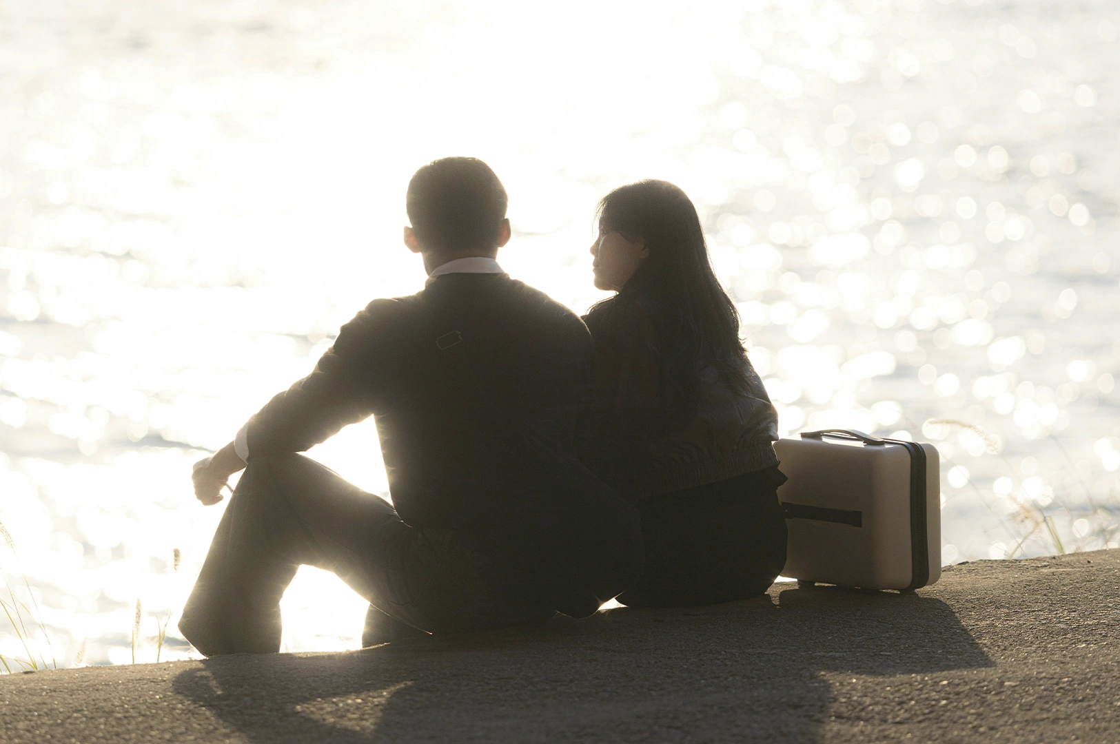 A poetic, back-lit silhouette shot of a couple sitting on a concrete ledge overlooking a shimmering body of water. The sun, positioned low in the sky, creates a brilliant white glare and a "bokeh" sparkling effect on the water's surface, washing the couple in a warm, ethereal glow. The man is on the left, and the woman sits next to him with a white hard-shell suitcase resting by her side. The overall mood is peaceful and reflective, capturing a quiet moment of connection at the end of a journey or during a sunset transition.