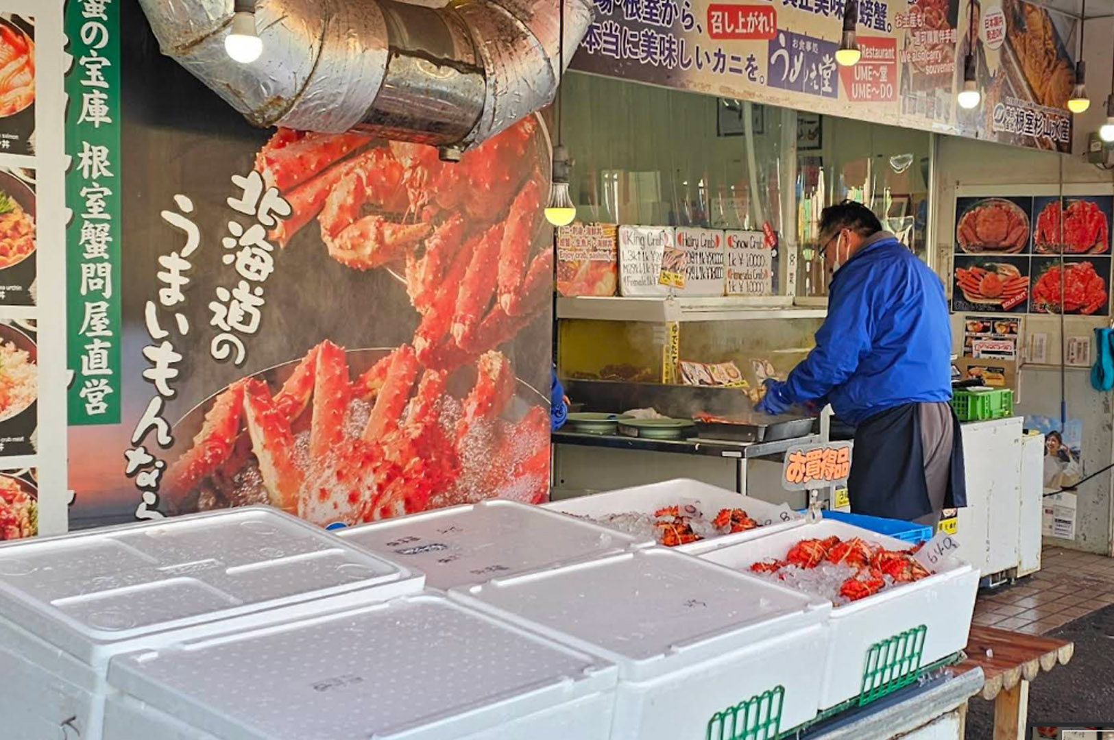 A close-up of a seafood stall specializing in Hokkaido crab. A large, realistic mural of a Red King Crab dominates the left side of the frame. In the foreground, white Styrofoam boxes filled with ice and fresh orange crabs are displayed for sale. To the right, a vendor in a blue jacket and apron works behind a steaming counter, with handwritten price signs for King, Hairy, and Snow crabs visible in the background.
