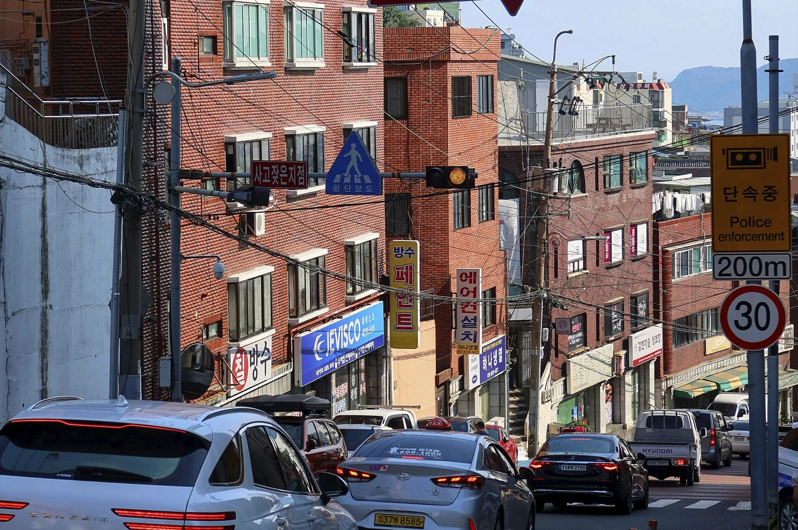 A view of a steep city street filled with traffic. In the foreground, the rear of a white Genesis SUV and a silver Hyundai Sonata are prominent. The street is lined with red-brick buildings featuring various commercial signs and a blue pedestrian crossing sign. A yellow "Police Enforcement" sign with a 30 km/h speed limit is visible on a pole to the right. In the far distance, a glimpse of the blue sea and a misty coastline can be seen.