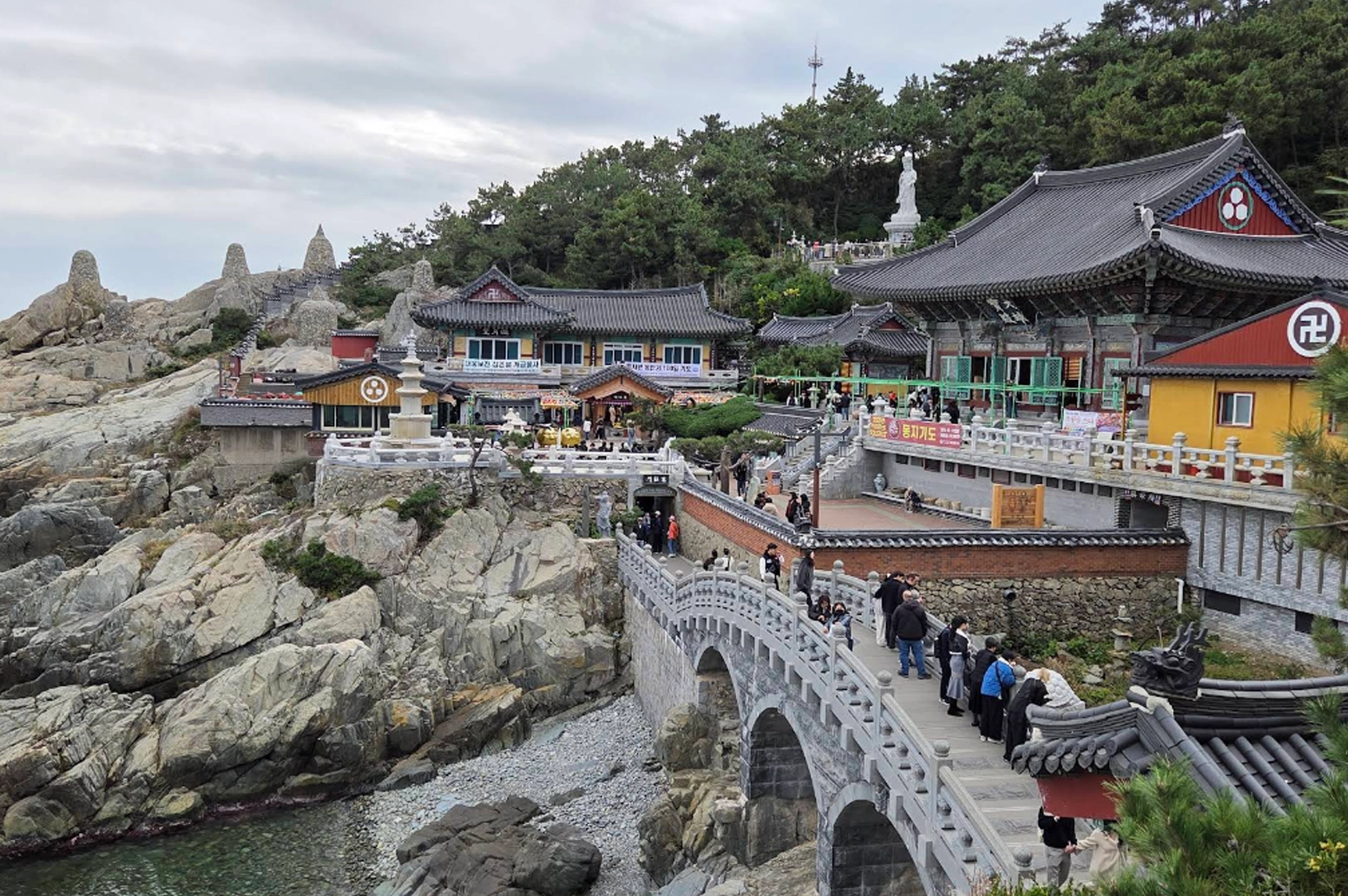 Haedong Yonggungsa Temple built directly into the rocky cliffs along the ocean. The image shows traditional Korean architecture with dark tiled roofs and colorful ornate details. A stone arched bridge is crowded with visitors crossing toward the main temple complex. In the background, a large white statue of Haesu Gwaneum Daebul (The Seawater Great Goddess of Buddha) stands among green pine trees.