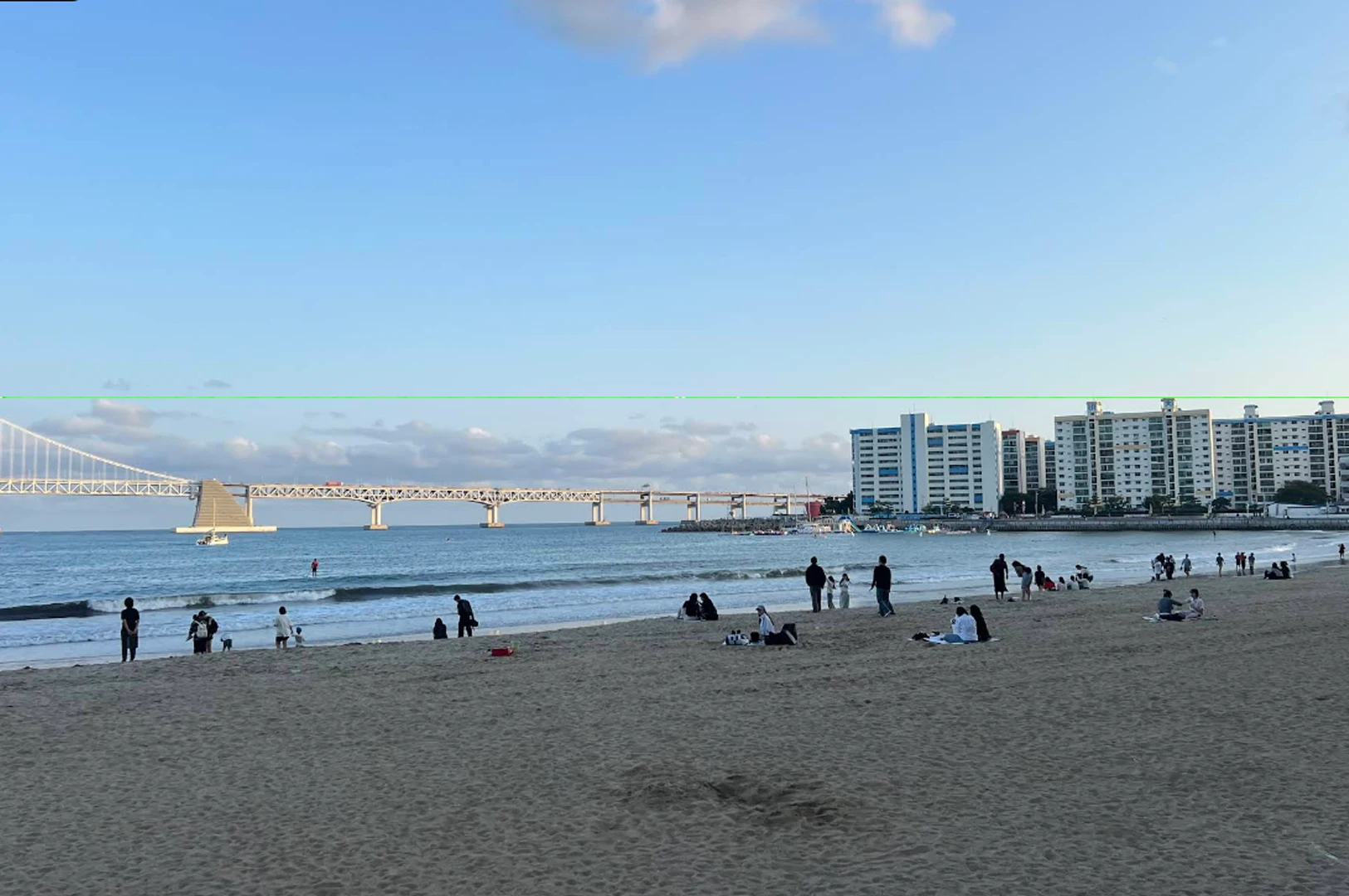 A scenic view of Gwangalli Beach at dusk. Numerous people are scattered across the wide sandy shore, some sitting on mats and others walking near the gentle surf. In the distance, the long, illuminated Gwangan Bridge (Diamond Bridge) stretches across the ocean. On the right side of the bay, several white and blue high-rise apartment buildings stand along the coast under a sky transitioning from blue to soft evening hues with scattered clouds.