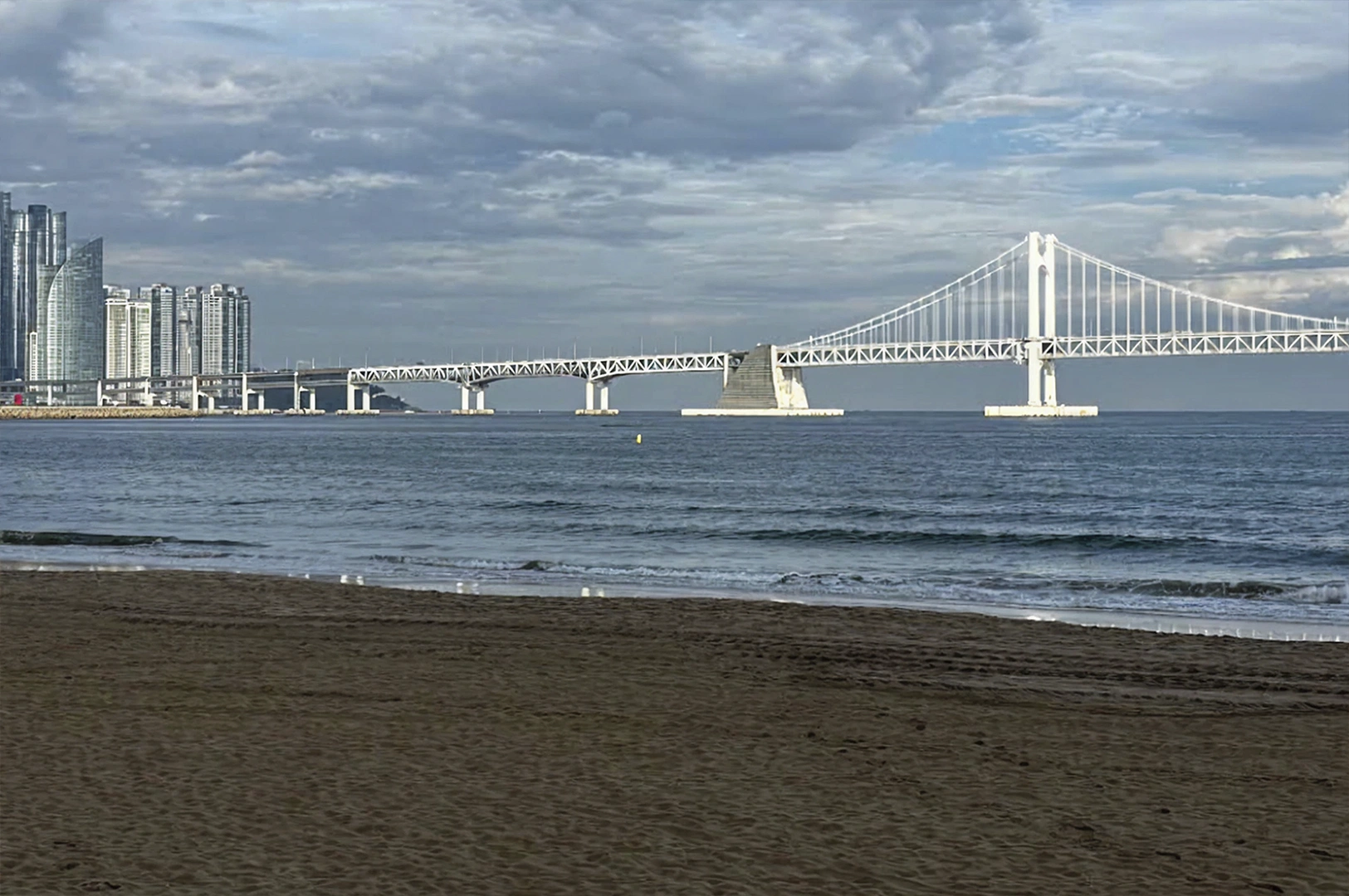 A low-angle view taken from the dark sands of Gwangalli Beach looking out toward the iconic Gwangan Bridge. The massive white suspension bridge stretches across the horizon, silhouetted against a moody, overcast sky filled with grey clouds. To the left, the modern skyscrapers of Marine City rise up from the water’s edge.