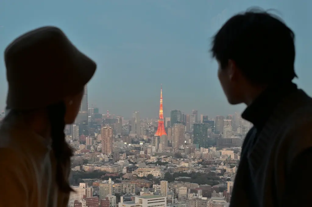 Two people are seen from behind in silhouette, looking out of a high-rise window at the sprawling Tokyo skyline during twilight. The iconic Tokyo Tower stands out in the center, illuminated in a vibrant orange glow against the blue and grey hues of the city’s dense buildings and skyscrapers. The person on the left wears a wide-brimmed hat, and both figures appear to be quietly taking in the expansive urban view as the sun sets.