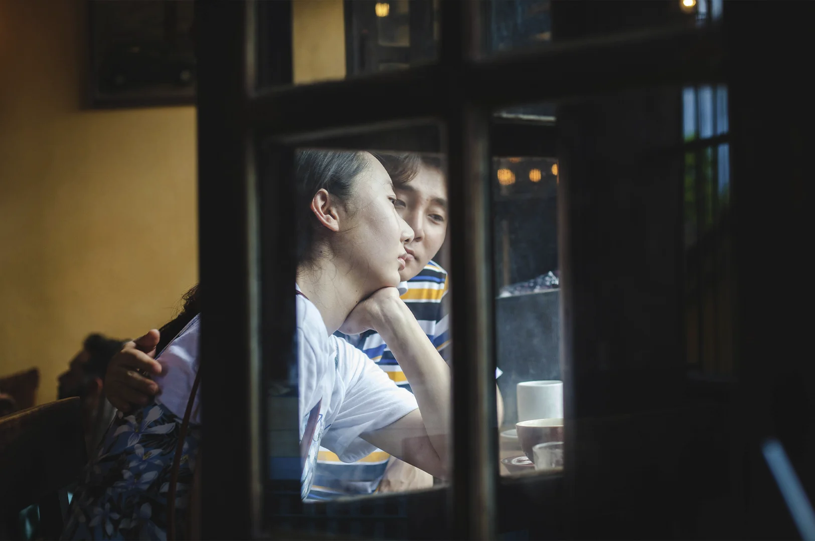 A voyeuristic, artistic shot taken through a dark wooden window frame, looking into a cozy, dimly lit café. A young woman with her hair pulled back rests her chin on her hand, looking thoughtfully into the distance with a melancholy or pensive expression. Beside her, a man in a yellow and blue striped shirt looks toward her with concern or affection. On the wooden table in front of them sits a white ceramic mug and a glass of water. The reflection on the window glass adds layers of depth, blending the interior intimacy with the outside world.