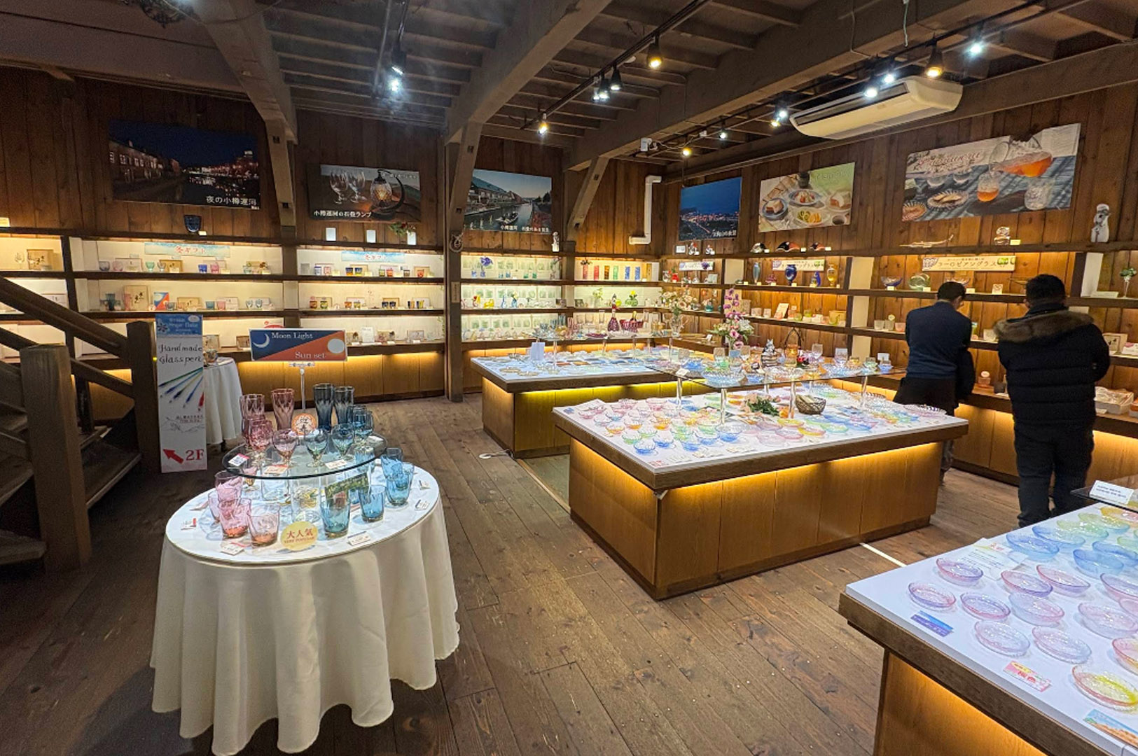 The interior of a warm, rustic shop featuring Otaru’s famous handmade glass. The room is characterized by dark wood floors, exposed ceiling beams, and wooden shelving units lining the walls. Glowing display tables in the center are covered in hundreds of colorful glass bowls and plates in pastel shades. Framed photos of Otaru scenery hang high on the walls.