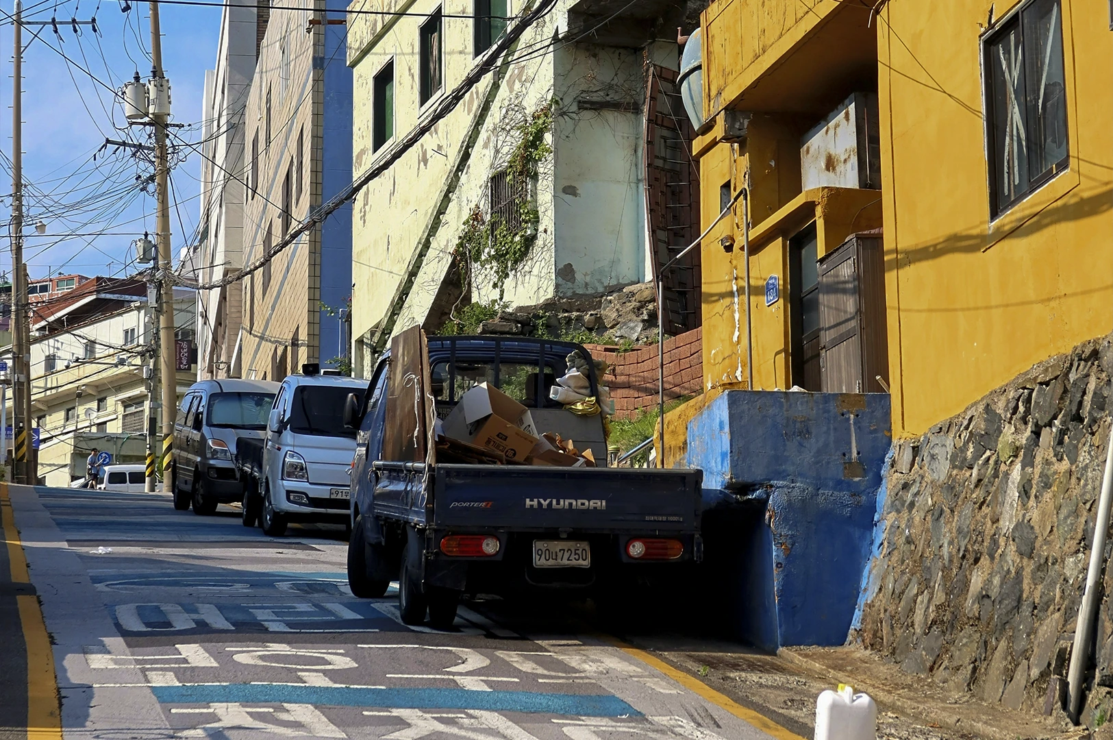 A steep, narrow side street with colorful buildings. A small blue Hyundai Porter pickup truck, loaded with flattened cardboard boxes, is parked on the left side of the incline next to a pale green building. On the right, a bright yellow building sits atop a stone retaining wall. Large white and blue Korean characters are painted directly onto the asphalt of the road. Overhead, a dense network of electrical wires crisscrosses against the blue sky.