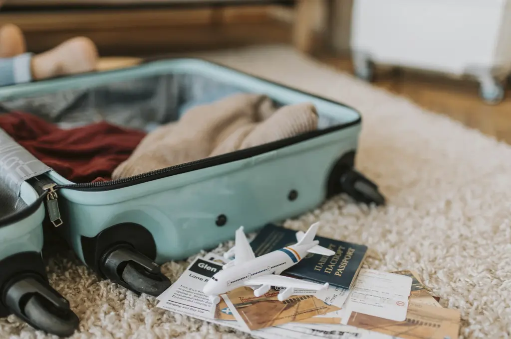A teal-colored hardshell suitcase lies open on a plush, cream-colored shag rug, partially packed with neutral-toned clothing. In the foreground, a small white toy airplane rests on top of a collection of travel documents, including a passport and several printed boarding passes or vouchers. The soft, warm lighting and shallow focus evoke a sense of anticipation and the final stages of preparation before embarking on a long journey.