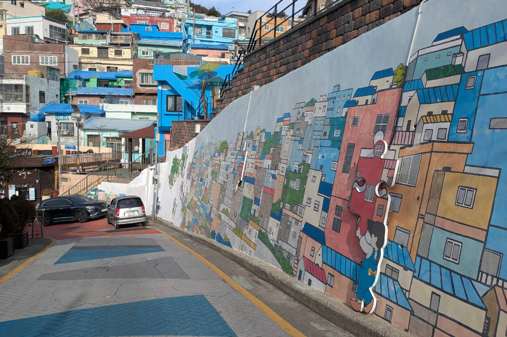 Colorful hillside in Gamcheon Culture Village. A large white retaining wall in the foreground features a vibrant mural of a boy in blue overalls holding a giant red puzzle piece, set against a backdrop of painted 2D houses. In the distance, real terraced houses painted in bright shades of blue, pink, and yellow climb the steep hill under a clear sky.