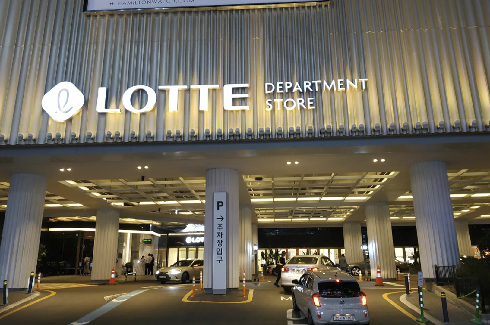 A night-time architectural shot of the entrance to a Lotte Department Store. The building's facade is made of vertical golden-lit fins with a large, glowing white "LOTTE DEPARTMENT STORE" sign. Below the overhang, bright recessed square lights illuminate a driveway where several cars are parked or moving. Large white circular pillars support the structure, and orange traffic cones and green poles line the asphalt path.