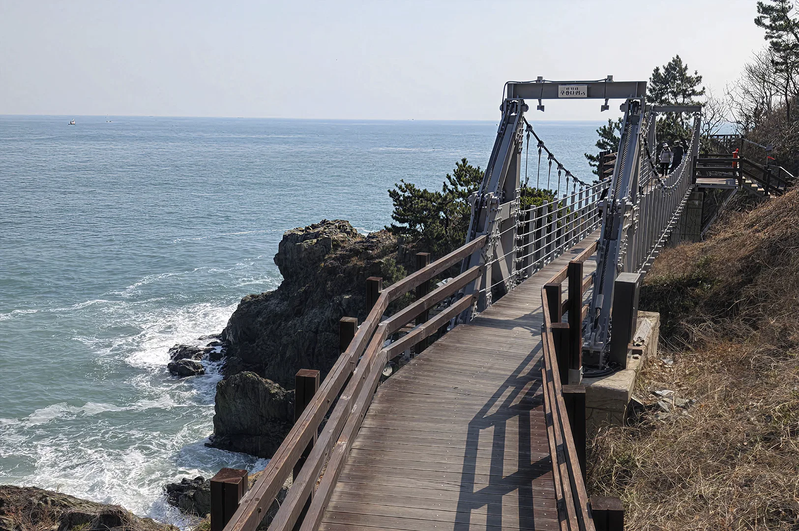 A first-person perspective looking down a wooden boardwalk and suspension bridge that hugs the side of a steep, rocky cliff. The bridge features metal railings and grey support structures. To the left, the turquoise sea surges against dark, jagged rock formations. Two hikers are seen further down the path, which leads toward a horizon where the water meets a pale, clear sky.
