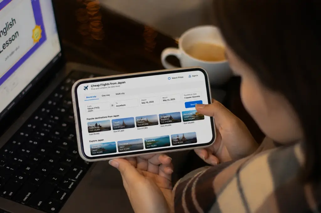 A person holds a large smartphone horizontally, showing a detailed mobile version of the Google Flights website. The screen displays a search for "Cheap Flights from Japan" with options for round-trip, one-way, and multi-city travel, showing specific dates in May 2025. Below the search bars, colorful thumbnails suggest popular destinations like Seoul, Taipei, and Bangkok. In the blurred background, a laptop screen and a white mug of coffee on a dark table suggest a cozy indoor environment for travel browsing.