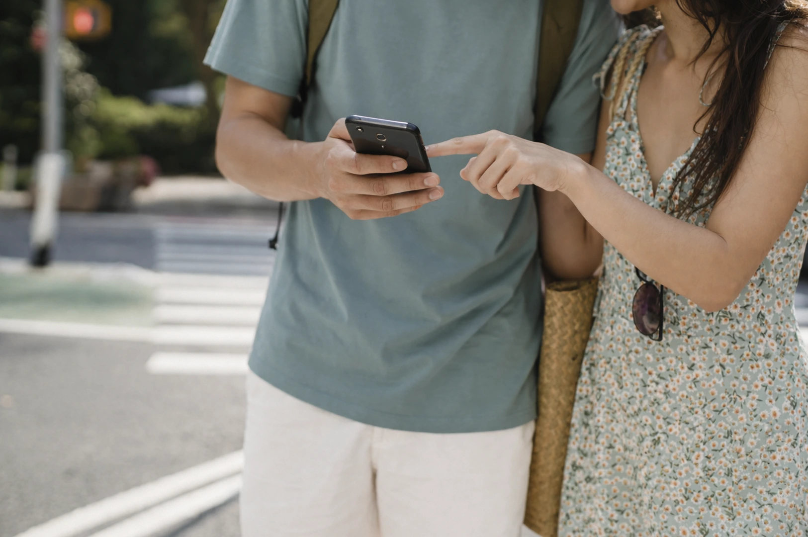 A close-up, waist-up shot of a couple standing on a sunny city street, focused on their interaction with a smartphone. The man, wearing a muted teal t-shirt and white shorts with a backpack strap visible, holds a black smartphone. The woman, dressed in a delicate green floral sundress with sunglasses tucked into her neckline, points at the screen with her index finger as they navigate their route. A straw tote bag hangs from her shoulder, and a blurred crosswalk and street signal in the background emphasize their status as tourists exploring a new urban environment.