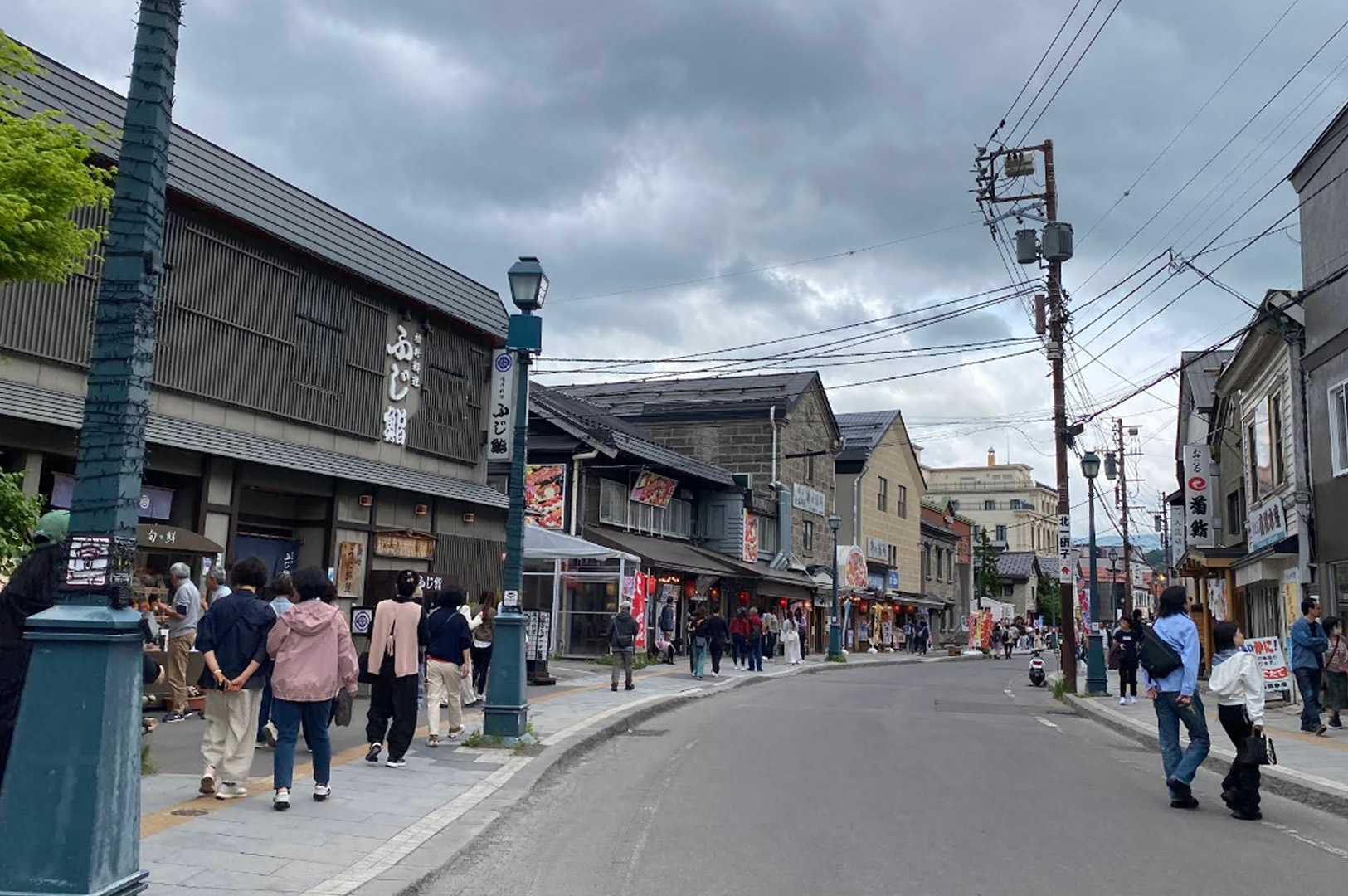 A view looking down Sakaimachi Street, a popular historic shopping district. The road curves gently past traditional stone and wood-panelled buildings housing sushi restaurants and boutiques. People in casual clothing stroll along the wide sidewalks. Green vintage-style lampposts and a dense network of overhead power lines add to the characteristic Japanese urban aesthetic under a cloudy sky.