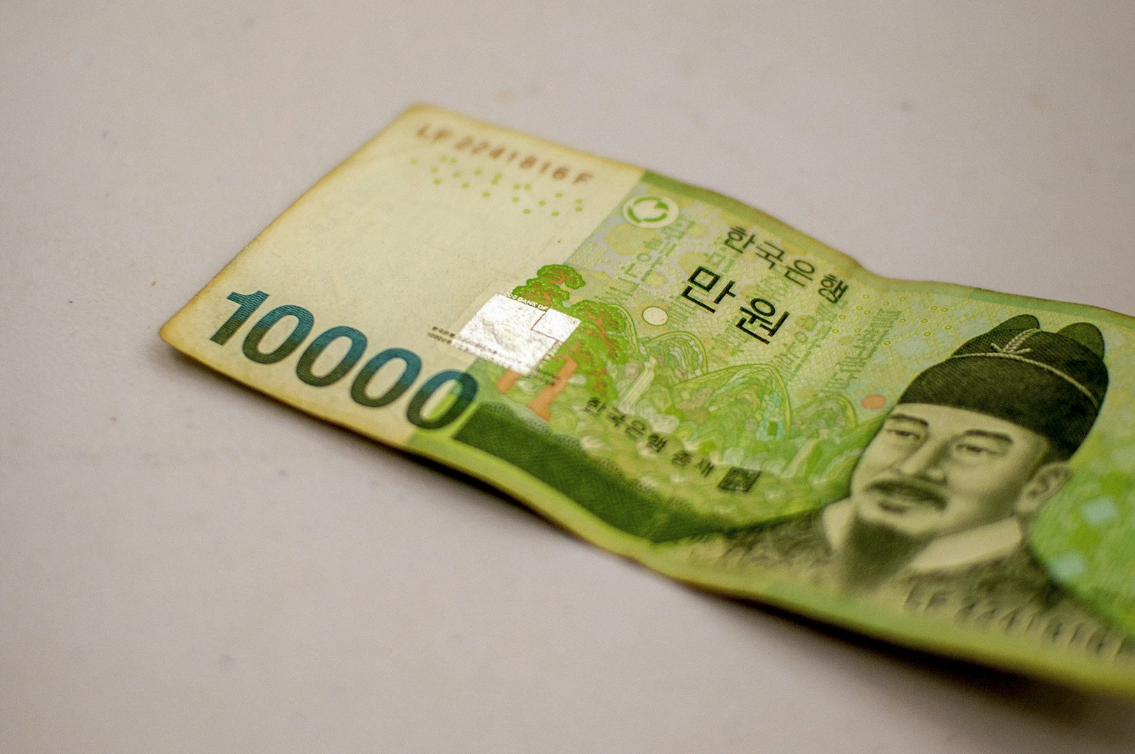 A high-angle, close-up shot of a single South Korean 10,000 won banknote resting on a neutral, off-white surface. The bill is slightly crumpled. The large green number "10000" is prominent on the left, while the right side features the portrait of King Sejong the Great. A small, square holographic security patch reflects light in the center.