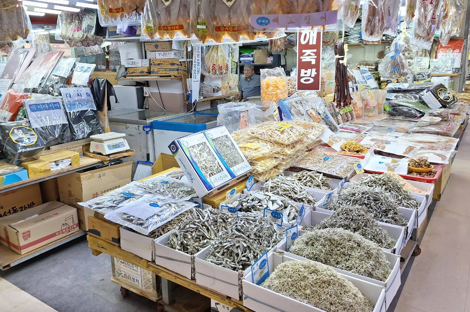 Bustling market stall filled with various dried seafood products. In the foreground, several white boxes are overflowing with different sizes of dried anchovies and small fish. Above the counter, clear plastic bags of dried squid and fish fillets hang from the ceiling. An elderly shopkeeper sits in the background behind stacks of packaged goods and colorful signage in Korean.
