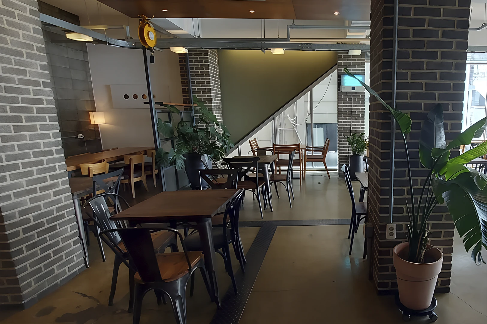 An interior view of a modern cafe featuring an industrial aesthetic. The space is characterized by exposed grey and brown brick pillars, concrete flooring, and an open ceiling with visible metal pipes and a yellow hoist pulley. Dark wooden tables are paired with a mix of black metal and light wood chairs. Potted green plants, including a large Monstera and a Bird of Paradise, add a natural element to the warm, moody lighting provided by a small desk lamp and recessed ceiling fixtures.