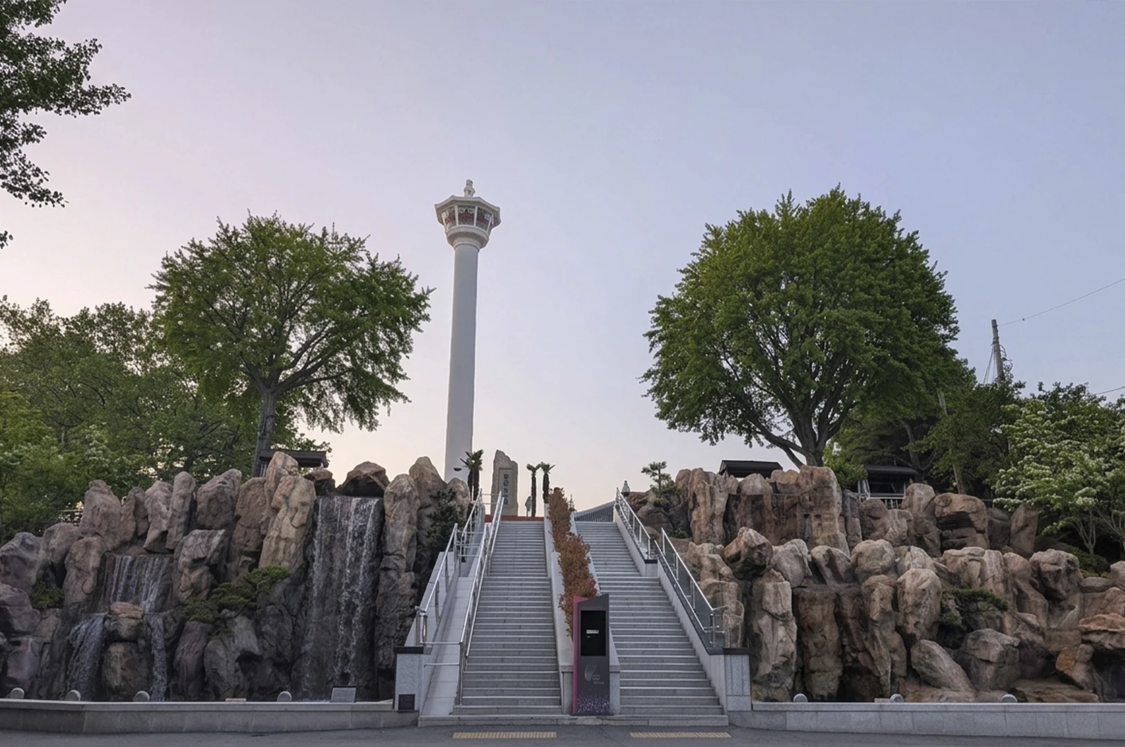A symmetrical shot of the white, slender Busan Tower rising into a soft, dawn-lit lavender sky. In the foreground, a wide stone staircase leads upward, flanked by artificial rock formations and a tiered waterfall. Large, manicured green trees frame the tower on both sides, creating a serene urban park setting at Yongdusan Park.