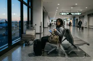 A woman with messy hair sits alone on a row of airport terminal seats during the evening or night. She wears a grey hoodie and looks tired, holding a smartphone in her hand. A large olive-green backpack and a black suitcase are next to her. The terminal is mostly empty, with large windows showing a dark blue sky and airport ground equipment outside.